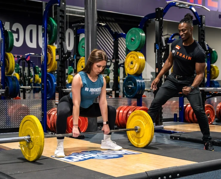 woman picks a barbell up off the ground next to her personal trainer